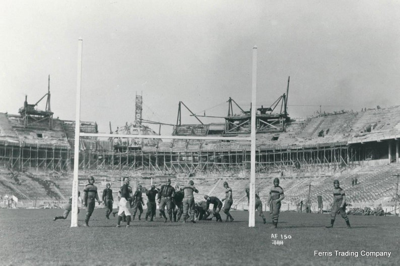 May include: A black and white photo of a football game being played in a stadium under construction. The players are wearing uniforms and are running with the ball. The stadium is in the background and is made of wood and metal.