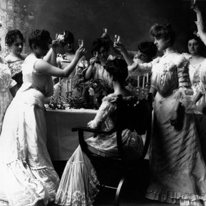 May include: A group of women in white dresses are gathered around a table, toasting with glasses of champagne. The women are smiling and laughing, and the atmosphere is festive. The table is decorated with candles and flowers. The image is in black and white, and the women are dressed in the style of the early 1900s. The text at the bottom of the image reads 