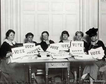 Op de afbeelding: Zeven vrouwen in zwart-witte kleding houden borden vast met de tekst "VOTE" in een kamer met een houten deur en een tafel. De vrouwen zitten aan de tafel en glimlachen.