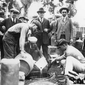 May include: A group of people standing around a barrel, pouring liquid from a jug into the barrel. There are several barrels and jugs in the background.