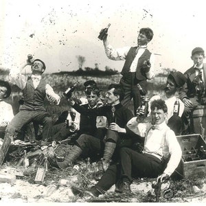 May include: A group of men in black and white clothing are gathered together outdoors. They are holding bottles and glasses and appear to be celebrating. There are several empty bottles on the ground and a crate of bottles in the background.
