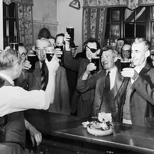 May include: A group of men in a pub, raising their glasses of beer in a toast. The men are wearing suits and ties, and they are all smiling. The pub is dimly lit, and there is a wooden bar in the background.