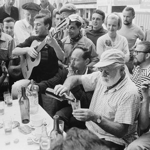 May include: A group of people gathered around a table, some are drinking from glasses, one person is pouring a bottle of wine into a glass, and another person is playing a guitar.