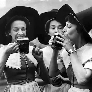 May include: Three women wearing black hats with wide brims and white lace collars are drinking from mugs. The women are smiling and appear to be enjoying themselves.