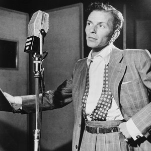 May include: A black and white photo of a man singing into a microphone. He is wearing a suit with a patterned tie and suspenders. The microphone is on a stand.