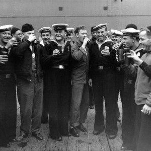 May include: A group of men in black and white sailor uniforms are standing on a deck. They are holding mugs and smiling. The men are wearing white hats with black bands. The men are standing in front of a gray wall.
