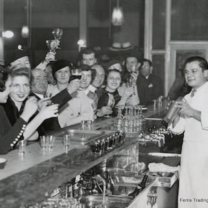 May include: A black and white photo of a group of people at a bar. The people are all smiling and raising their glasses in a toast. A bartender is shaking a cocktail shaker behind the bar. The text 