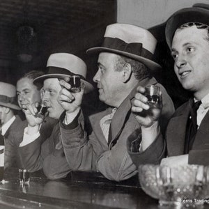 May include: Four men sitting at a bar, each holding a glass of liquor. The man on the far right is wearing a dark suit and a fedora. The man next to him is wearing a light suit and a fedora. The man in the middle is wearing a dark suit and a fedora. The man on the left is wearing a light suit and a fedora. The bar is dark and there are bottles of liquor on the counter. The text 