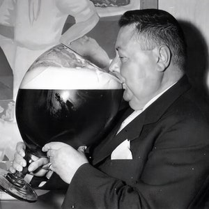 May include: A large, stemmed glass brimming with a dark liquid, possibly beer, topped with a thick head of foam. The glass is substantial, with a wide bowl and a sturdy base. The liquid appears dark and rich, suggesting a robust beverage. The glass is presented in a monochrome palette, enhancing the classic feel of the image.