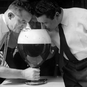 May include: Two people are leaning over a large glass of beer with a thick head of foam. The glass is on a table and the people are looking at the beer.
