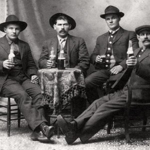 May include: Four men in suits and hats are sitting around a table. They are holding bottles of beer and glasses. The table has a floral tablecloth. The men are smiling and looking at the camera. The image is in black and white. The text 