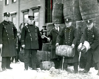 May include: Black and white photo of several men in uniform pouring liquid from barrels into the snow. The men are wearing hats and coats. There are several barrels in the background.