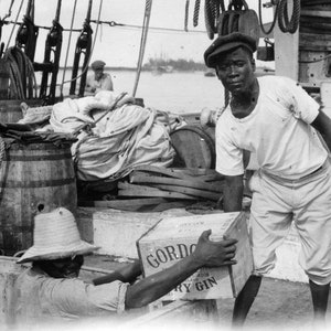 May include: A black and white photo of two men on a ship. One man is wearing a white shirt and white pants. He is holding a box that says 