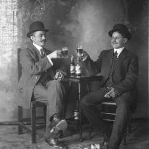 May include: Two men in suits and hats are sitting at a table, each holding a glass of beer and toasting each other. There are two bottles of beer on the table.
