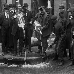 May include: A group of men in black and white clothing standing in a street. Some of the men are pouring liquid from a pitcher into a puddle on the ground. The liquid is flowing through the puddle and creating a stream of water.