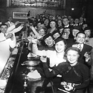 May include: A black and white photo of a group of people gathered around a bar. The people are all dressed in formal attire and are holding drinks. The bar is decorated with a variety of bottles and glasses. The people are smiling and laughing. The photo is taken from a low angle, looking up at the people.