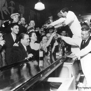 May include: A black and white photo of a group of people at a bar. The bartender is pouring a drink for a customer. The customers are all smiling and laughing. There is a piano in the background. The text 