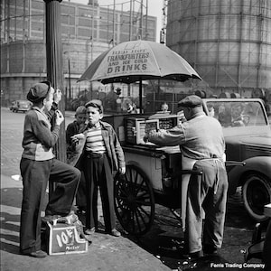 May include: A black and white photo of a street vendor selling red hot frankfurters and ice cold drinks from a cart. The vendor is wearing a gray sweater and a cap. Three boys are standing in front of the cart. The cart has a sign that reads 