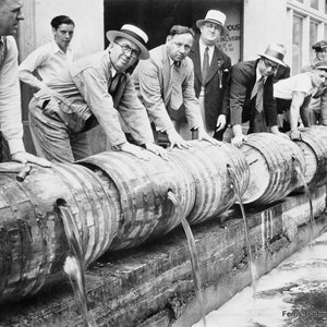 May include: A group of men in suits and hats are standing around a row of wooden barrels, pouring liquid from the barrels. The barrels are lined up on a concrete surface. The text 