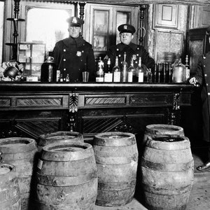 May include: Three police officers stand in a bar with a counter and several barrels in the foreground. The officers are wearing dark blue uniforms and hats. The counter is made of dark wood and has a variety of bottles and glasses on it. The barrels are made of wood and are stacked on top of each other.