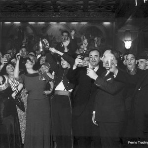 May include: A black and white photo of a group of people celebrating. The people are all dressed in formal attire and are holding glasses of champagne. They are all smiling and laughing. The photo is taken in a dimly lit room with a large painting on the wall behind them. The text 