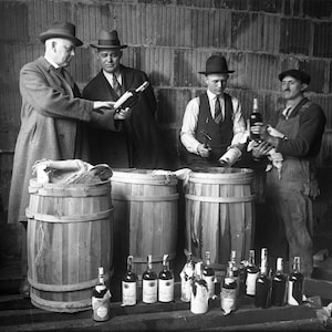 May include: Four men stand in front of three wooden barrels. The men are wearing hats and coats. There are several bottles of alcohol in front of the barrels.