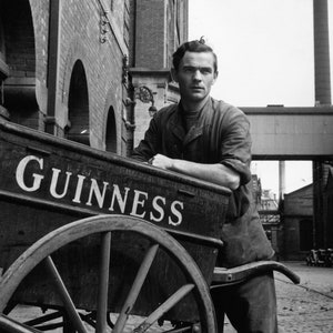 May include: A black and white photo of a man standing next to a wooden cart with a white 