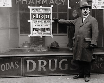 May include: A black and white photo of a storefront with a sign that reads "Public Notice These Premises Have Been Closed For One Year Violation Prohibition Law". A man in a coat and hat stands in front of the store, pointing to the sign.
