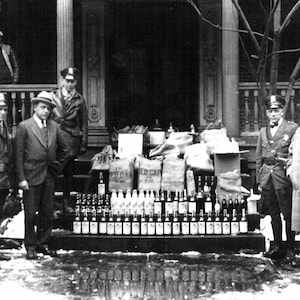 May include: A black and white photo of a group of people standing in front of a building. There are several bottles of alcohol stacked in front of them. The bottles are labeled with the words 