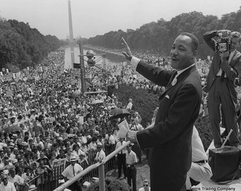 May include: A black and white photo of Martin Luther King Jr. giving a speech to a large crowd of people. The crowd is gathered in front of the Lincoln Memorial in Washington, D.C. King is wearing a suit and tie, and he is holding a microphone in his right hand. He is gesturing with his left hand. The crowd is cheering and waving their hands in the air.