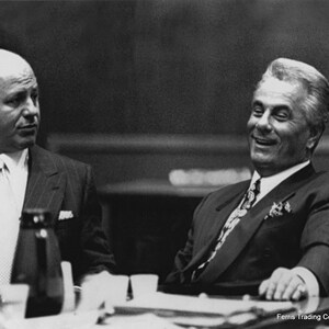 A vintage black and white photograph of notorious Gambino crime family boss John Gotti sitting at a table in a sharp suit and silk tie. He is laughing and leaning back confidently, capturing the charismatic yet dangerous persona of the Dapper Don