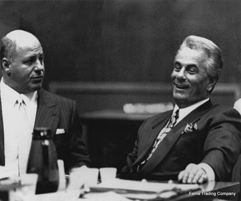 A vintage black and white photograph of notorious Gambino crime family boss John Gotti sitting at a table in a sharp suit and silk tie. He is laughing and leaning back confidently, capturing the charismatic yet dangerous persona of the Dapper Don