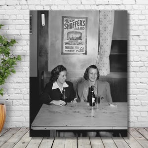 May include: Two women are sitting at a table in a restaurant. They are both smiling and looking at the camera. The woman on the left is wearing a dark jacket and a white shirt. The woman on the right is wearing a light jacket and a white shirt. There is a poster on the wall behind them that says 
