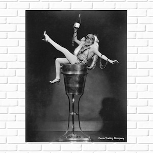 May include: A black and white photograph of a woman in a beaded dress sitting in a metal bucket on a stand. The woman is holding a bottle of champagne in her right hand and is looking at the camera. The photo is captioned 