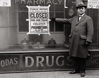 May include: A black and white photo of a storefront with a sign that reads "Closed for one year due to violation of the National Prohibition Law". A man in a coat and hat stands in front of the store, pointing to the sign.