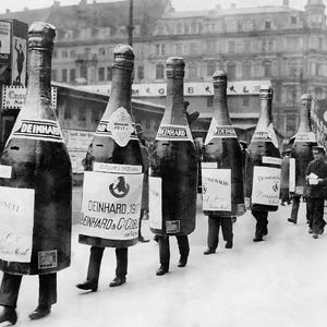 May include: A black and white photo of a group of people dressed as champagne bottles. The bottles are all wearing hats and carrying signs. The signs have the text 