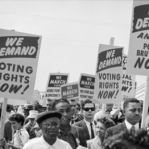 Op de afbeelding: Zwart-witfoto van een protestmars. Mensen dragen borden met slogans als "We Demand Voting Rights Now!" en "An End to Police Brutality Now!". Andere borden roepen op tot banen en eerlijk loon.