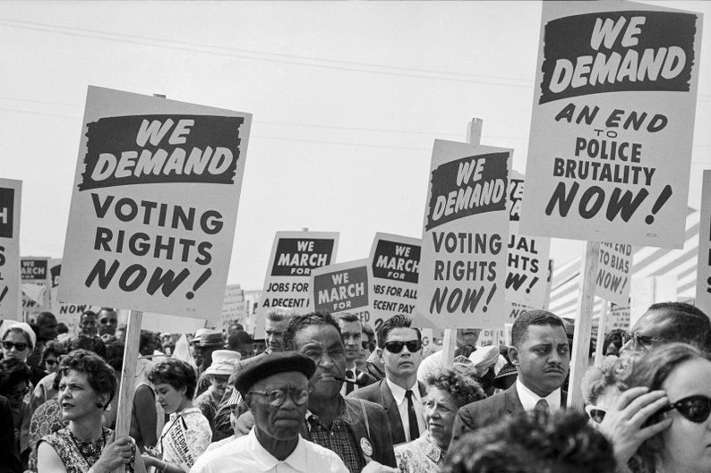 Op de afbeelding: Zwart-witfoto van een protestmars. Mensen dragen borden met slogans als "We Demand Voting Rights Now!" en "An End to Police Brutality Now!". Andere borden roepen op tot banen en eerlijk loon.