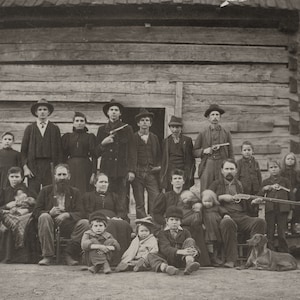 May include: Sepia-toned photograph of a large group of people posing in front of a log cabin. Many individuals are holding firearms, including rifles and pistols. The group includes men, women, and children, all dressed in period clothing.