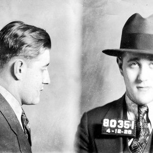 May include: Black and white mugshot of a man wearing a suit and a fedora. The mugshot shows both a profile view and a frontal view. The man has a number tag on his suit that reads "80351 4-12-28".