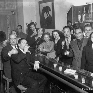 May include: A black and white photo of a group of people gathered around a bar. The people are all dressed in formal attire and are holding glasses of drinks. There are several glasses of drinks on the bar, and a pack of cigarettes is visible on the bar. The text 