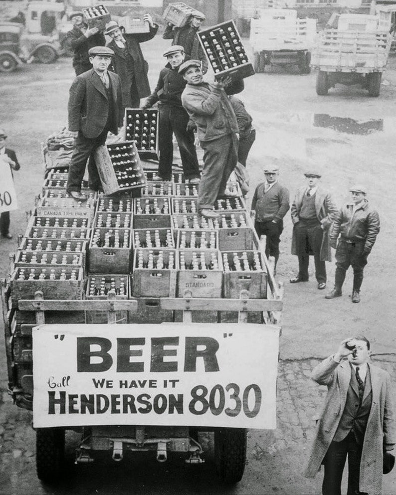 A vintage black and white photograph of men in flat caps and coats loading crates of beer onto a truck in Cleveland, Ohio. A large banner on the back of the truck reads "BEER" Call WE HAVE IT HENDERSON 8030.