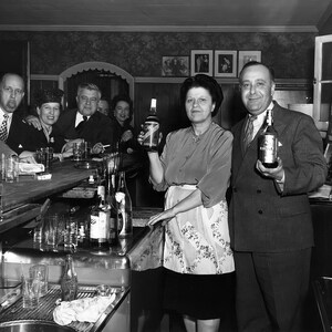 May include: A group of people standing at a bar counter. There are bottles of liquor on the counter and glasses in front of them. The people are all dressed in 1940s clothing. The bar is dark wood and has a backsplash of light colored tiles. There are several bottles of liquor on the counter, including a bottle of whiskey and a bottle of gin.