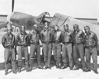 May include: A group of eight men in military uniforms stand in front of a black and white P-51 Mustang fighter plane. The men are wearing leather jackets, goggles, and helmets. The plane has the number "70" painted on the side.