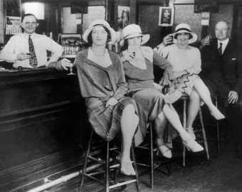 May include: Four people, two women and two men, are sitting at a bar. The women are wearing hats and dresses. The men are wearing suits. The bar is made of dark wood and has a counter with a black surface. There are glasses on the counter.
