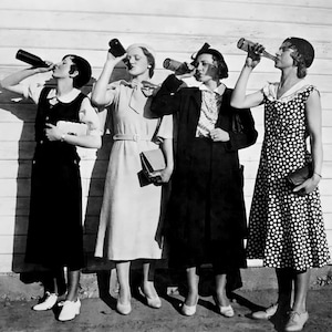 May include: Four women in 1920s clothing stand in front of a white wooden wall, each holding a bottle and drinking from it.