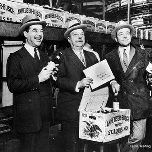 May include: Three men in suits and hats stand in front of a case of Anheuser-Busch beer. The case is labeled 