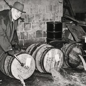 May include: A black and white photo of a man in a hat and coat standing next to a wooden barrel with a spigot. The barrel is labeled 