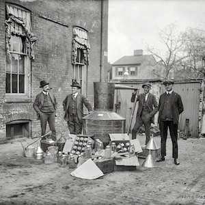 May include: A black and white photo of a group of men standing in front of a brick building. The men are wearing suits and hats. There is a large metal container in the center of the image, and a variety of glass jars and metal containers are scattered around it. The text 