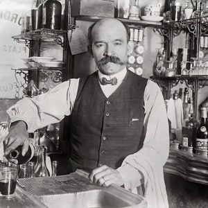 May include: A black and white photo of a man pouring a drink from a bottle into a glass. The man is wearing a vest and a bow tie. The background is a bar with shelves of bottles and glasses.